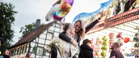 Eine junge Frau trägt eine andere junge Frau auf den Schultern, sie gehen fröhlich über eine Kirmes.