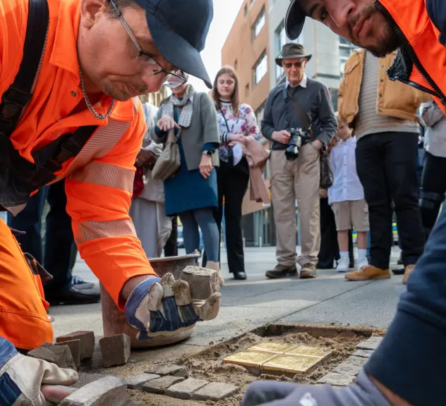 Im Vordergrund zwei Männer, die die Stolpersteine verlegen, im Hintergrund weitere Personen.