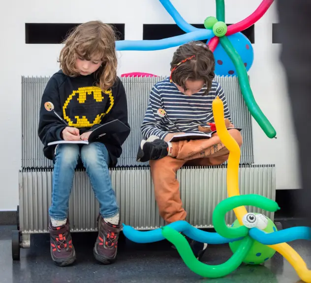 Zwei Kinder sitzen mit bunten Luftballons behangen auf einer Bank und schreiben auf Blöcke.