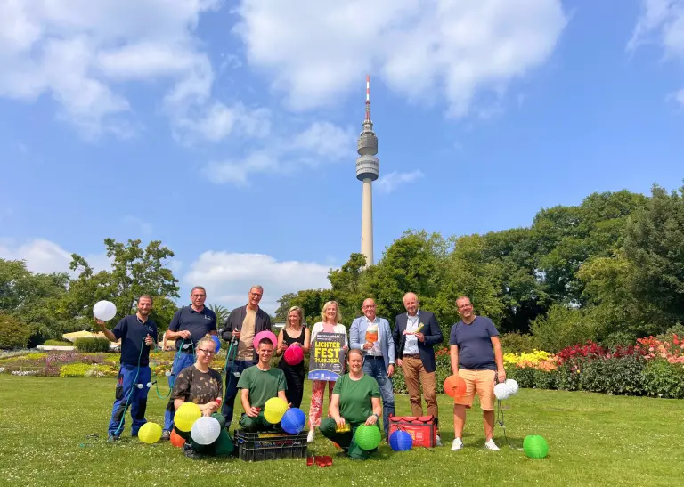 Eine Gruppe Menschen steht mit Lampions auf einer Wiese, dahinter ist der Florianturm zu sehen.