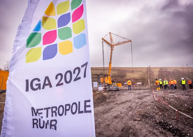 Eine Flagge der Internationalen Gartenausstellung 2027 im Vordergrund, im Hintergrund die Baustelle an der Kokerei Hansa.