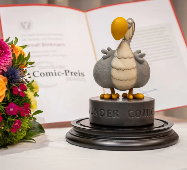 Der Dortmunder Comicpreis, ein Dodo im Comic-Stil, steht auf einem Tisch mit einem Blumenstrauß und der Urkunde dahinter