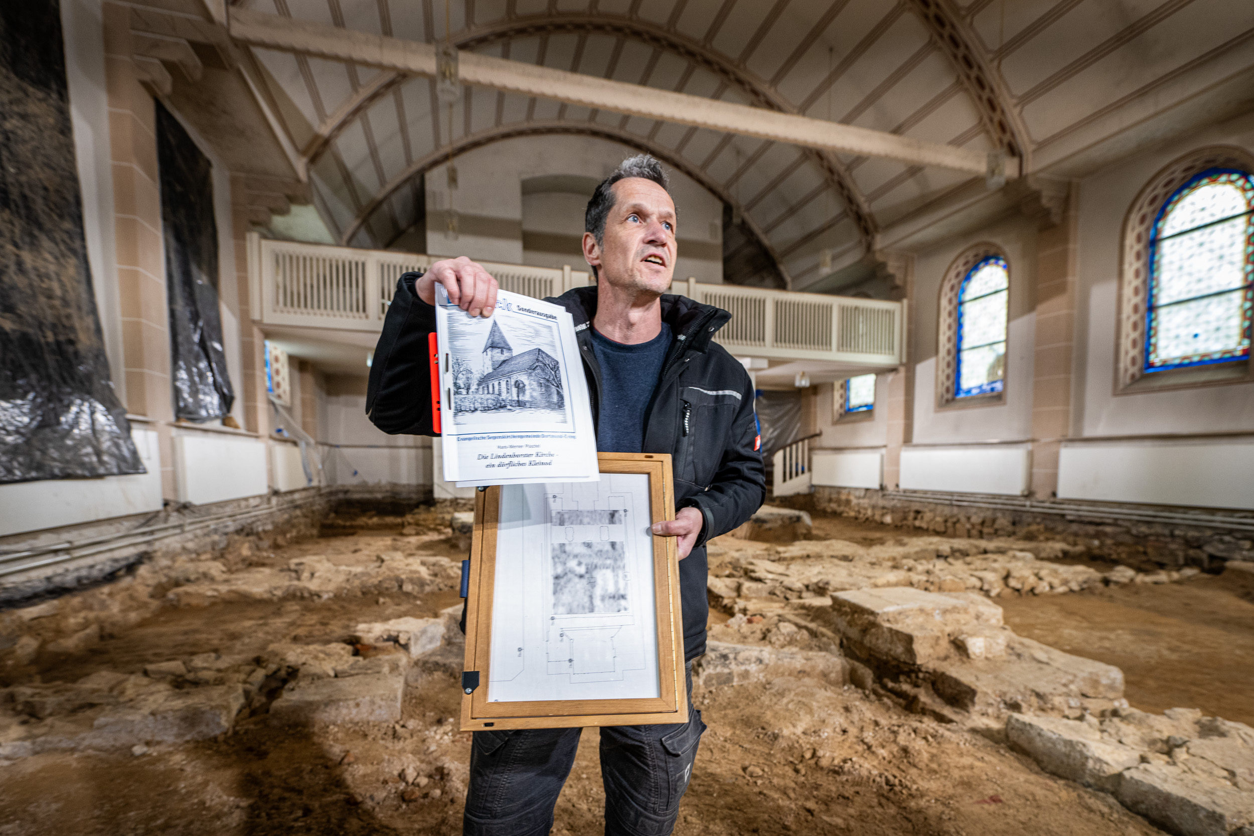 Ein Mann steht inmitten der archäologischen Funde unter der Lindenhorster Kirche in Dortmund und hält Dokumente hoch.