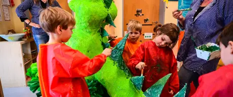 Drei Kinder malen einen Drachen aus Pappmaché grün an. Ein Erzieherin hilft ihnen.