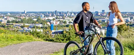 Zwei Jugendliche in der Natur vor der Stadt-Skyline. Einer hält ein Fahrrad.