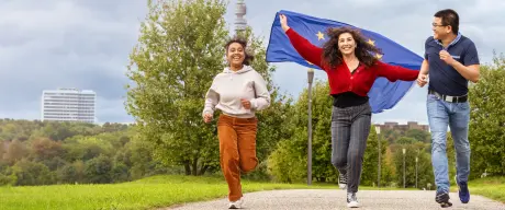 Drei Jugendliche rennen. Im Hintergrund ist der Florianturm zu sehen. Das Mädchen in der Mitte lässt eine Europa-Flagge hinter sich flattern, alle drei lachen. 