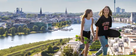 Zwei Mädchen spazieren mit ihren Skateboards unter dem Arm über einen Hügel, im Hintergrund ist der Phönixsee zu sehen