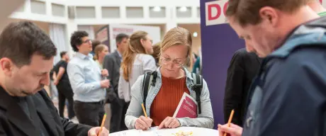 Drei Personen stehen an einem Stehtisch und schreiben auf Blätter.