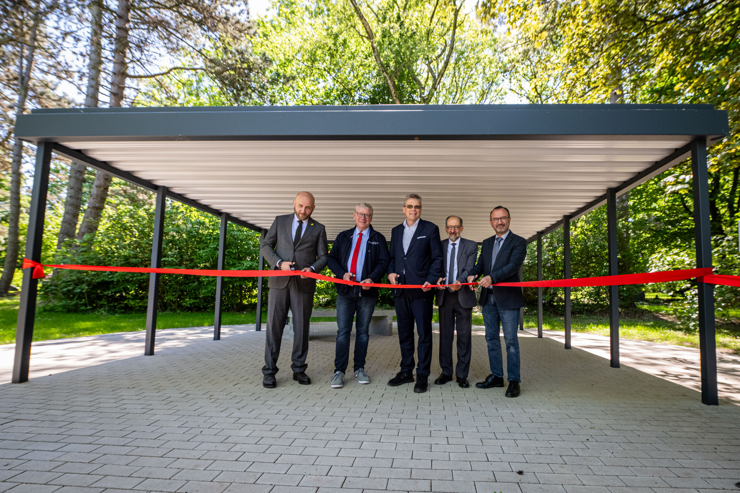 Fünf Männer schneiden symbolisch ein rotes Band vor einer Überdachung durch. Im Hintergrund zu sehen ist der Gebetstisch auf dem Gebetsplatz für Muslim*innen auf dem Hauptfriedhof.