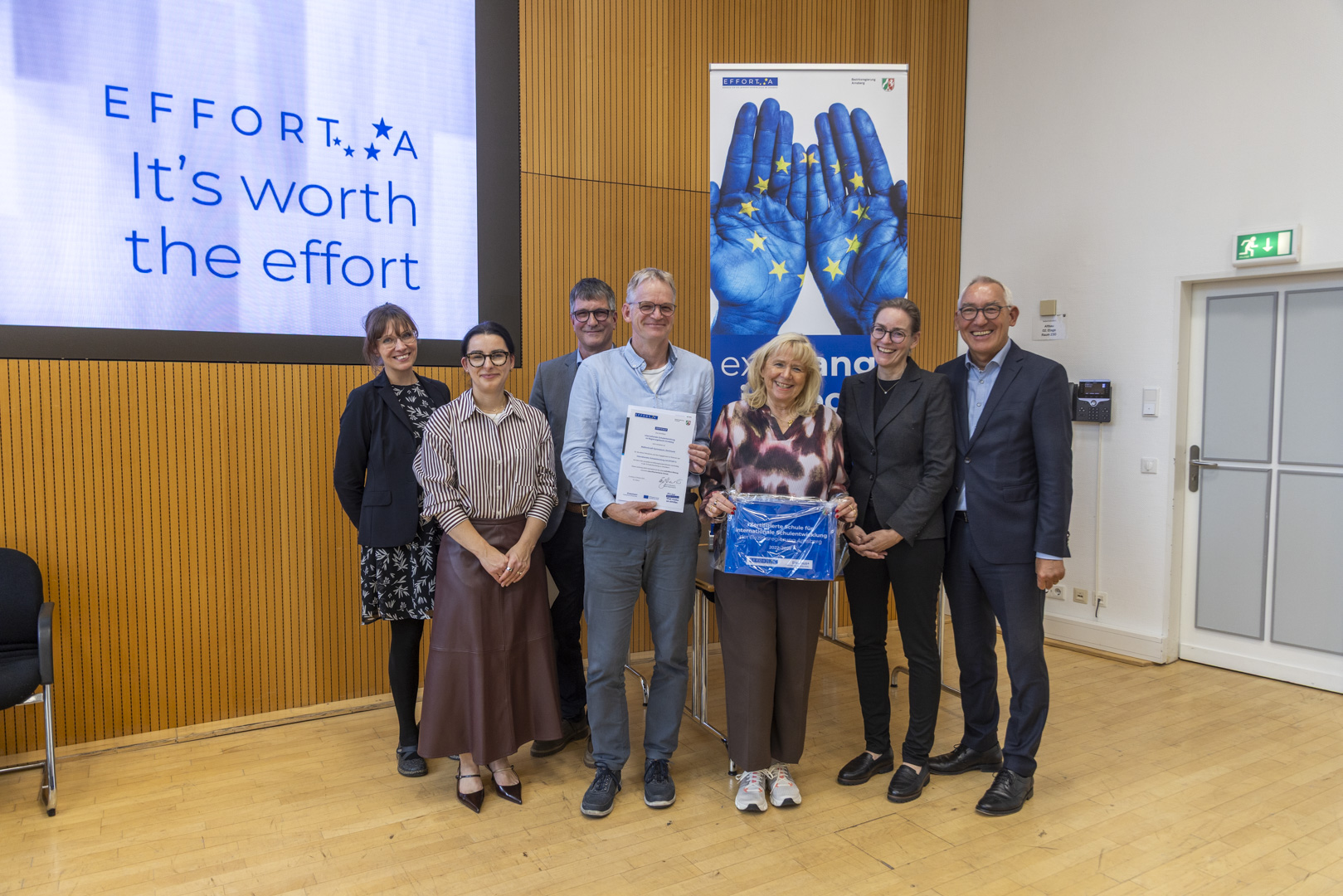 Eine Gruppe von sieben Personen steht in einem Saal vor einer Wand mit Holzverkleidung. Zwei Personen in der Mitte halten eine Urkunde und eine Auszeichnung. Im Hintergrund sind zwei Banner mit dem Schriftzug „EFFORT-A – It’s worth the effort“ und blauen, mit EU-Sternen bemalten Händen zu sehen.