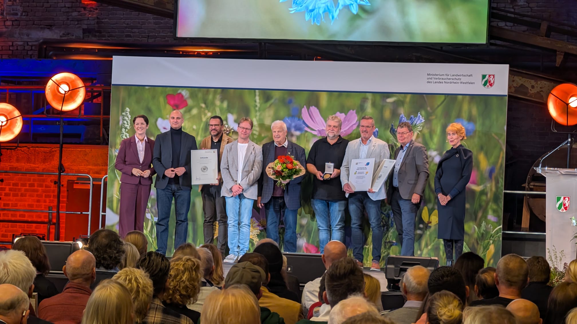 GruppenMehrere Personen stehen nebeneinander, einige halten Urkunden und Auszeichnungen sowie einen Blumenstrauß. Im Hintergrund ein großes Blumenmotiv, vor der Bühne sitzt Publikum.