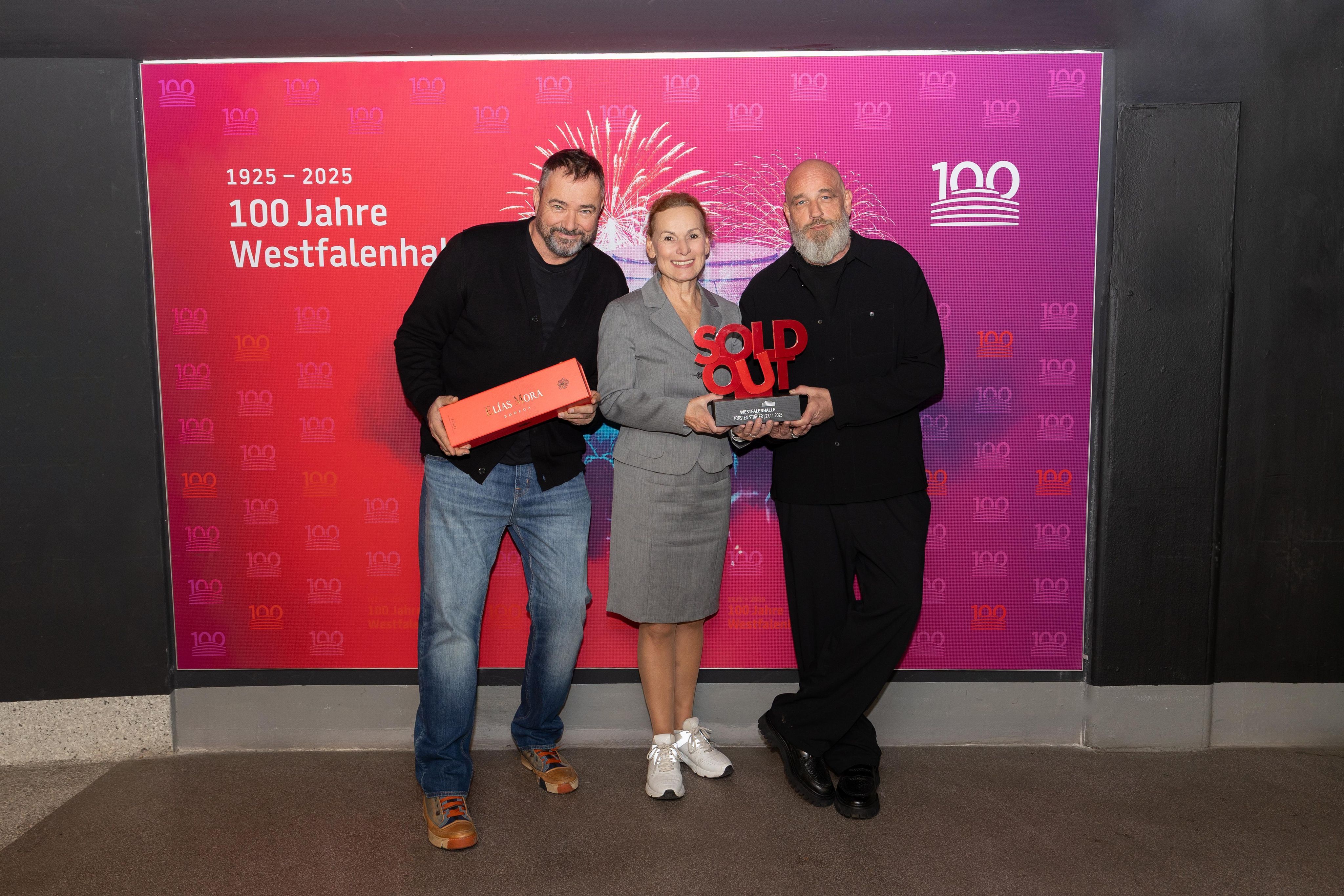 Udo Lühr, Sabine Loos und Torsten Sträter mit dem Sold-Out-Award der Westfalenhallen vor einer Fotowand.