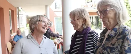 Drei ältere Frauen stehen auf einem Balkon und lachen. Dahinter sind noch mehr Menschen zu sehen.