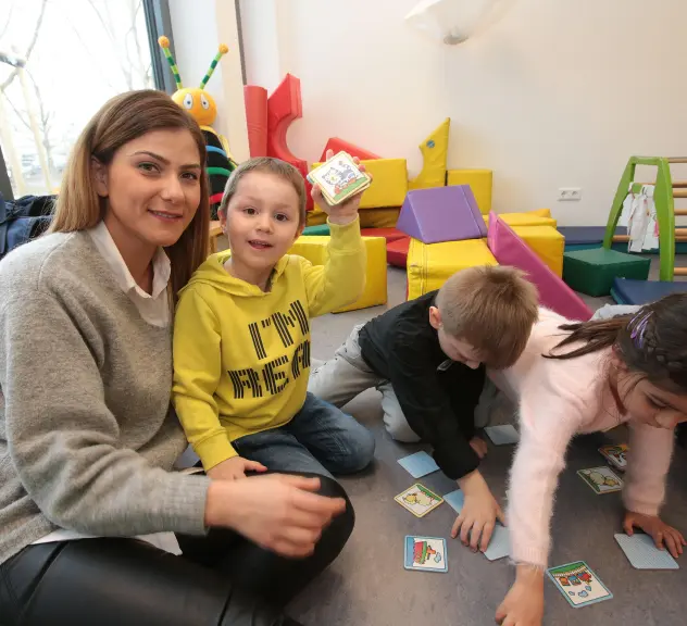 In einer Kita spielt eine Frau mit Kindern Memory und schaut für die Bildaufnahme in die Kamera.