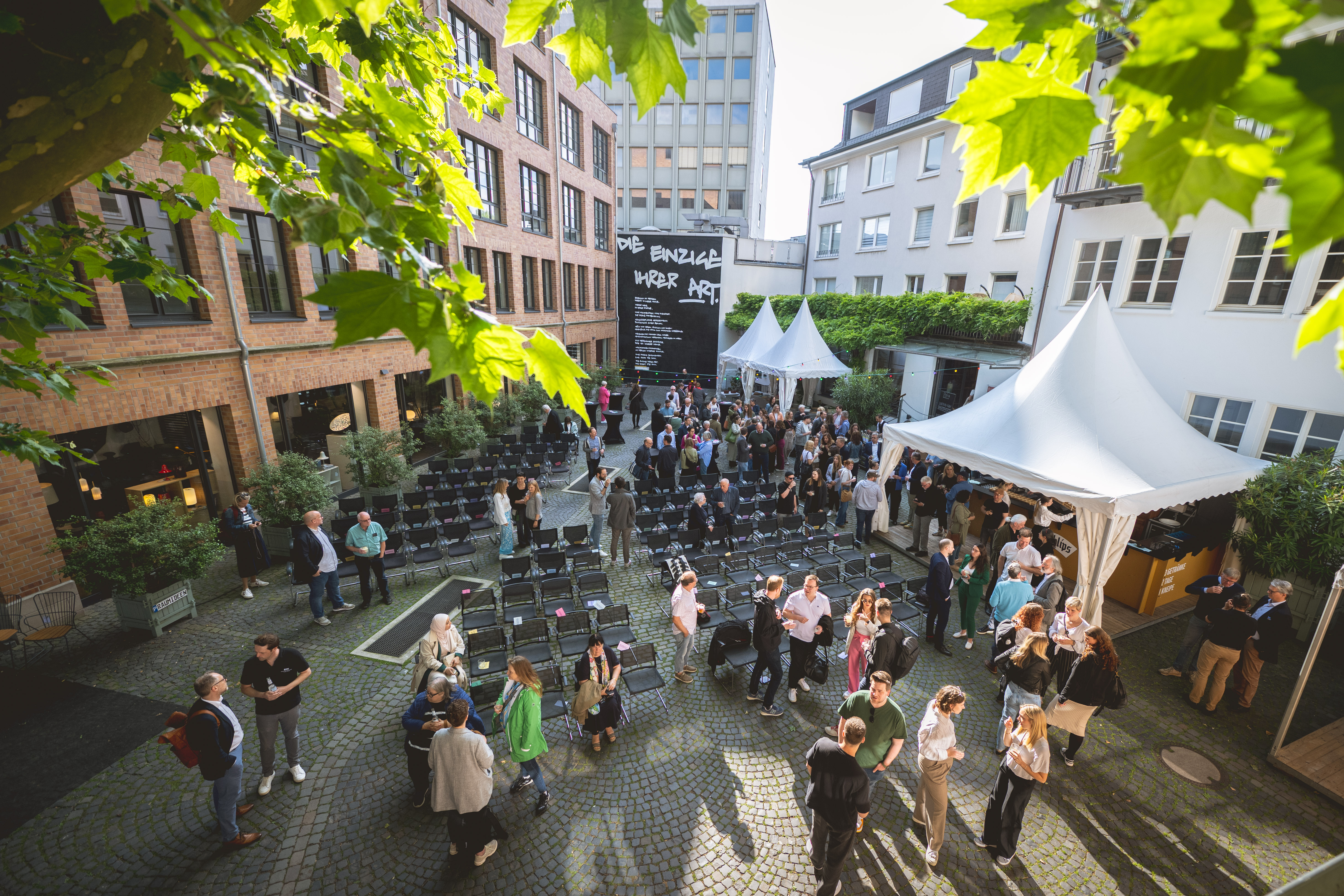 Ein Innenhof von oben. In der Mitte sind Stuhlreihen, darum verteilt sind einige Menschen im Gespräch miteinander. Es sind Zelte zu sehen für Getränke und Essen. Die Sonne scheint.