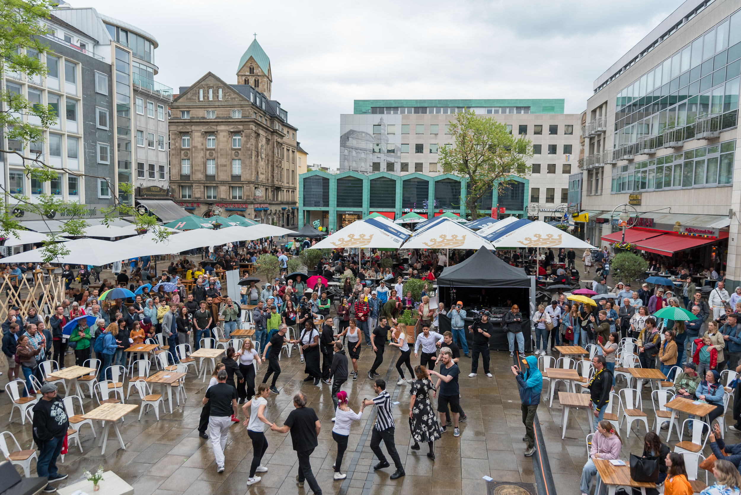 Eine Menschenmenge tanzt auf dem Marktplatz. Drumherum Zuschauende, Sonnenschirme und Bühnen.