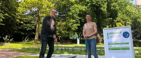 Zwei Personen spielen Minigolf im Westfalenpark Dortmund, umgeben von grünen Bäumen und dem Fernsehturm im Hintergrund.