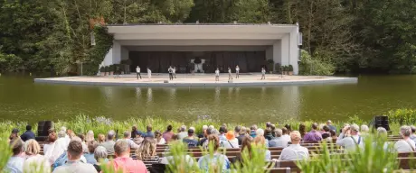 Die Seebühne am Buschmühlenteich im Westfalenpark während einer Sonntagsveranstaltung, mit Publikum und einem Ensemble auf der Bühne vor grüner Waldkulisse.