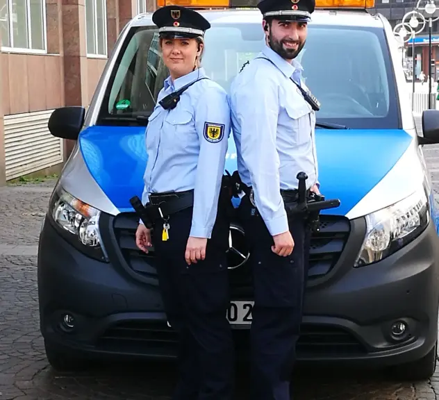 Zwei Auszubildende des Kommunalen Ordnungsdienstes stehen Rücken an Rücken vor einem Fahrzeug des Ordnungsamtes.