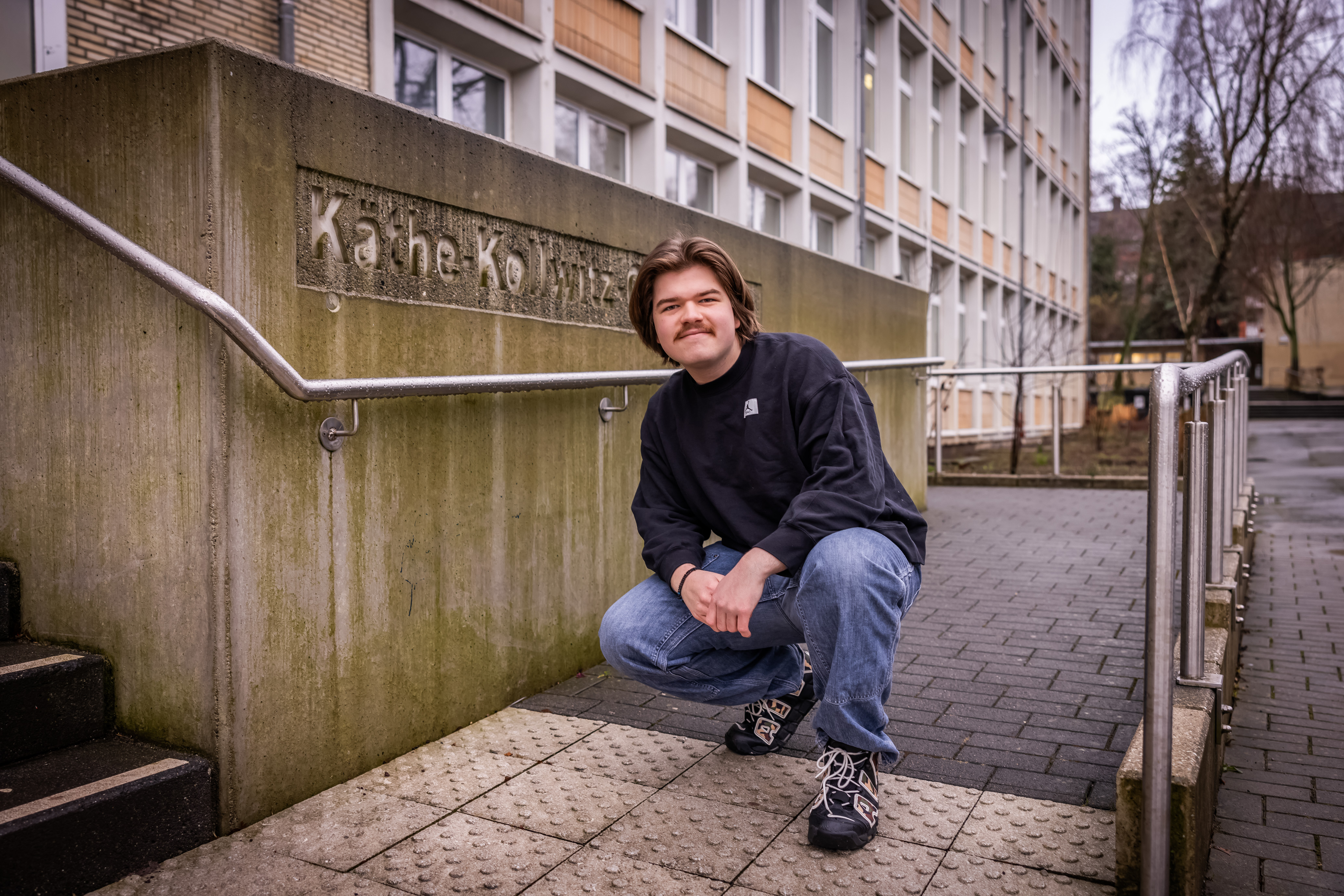 Bild eines jungen Mannes vor einem Schild mit der Aufschrift Käthe-Kollwitz-Gymnasium