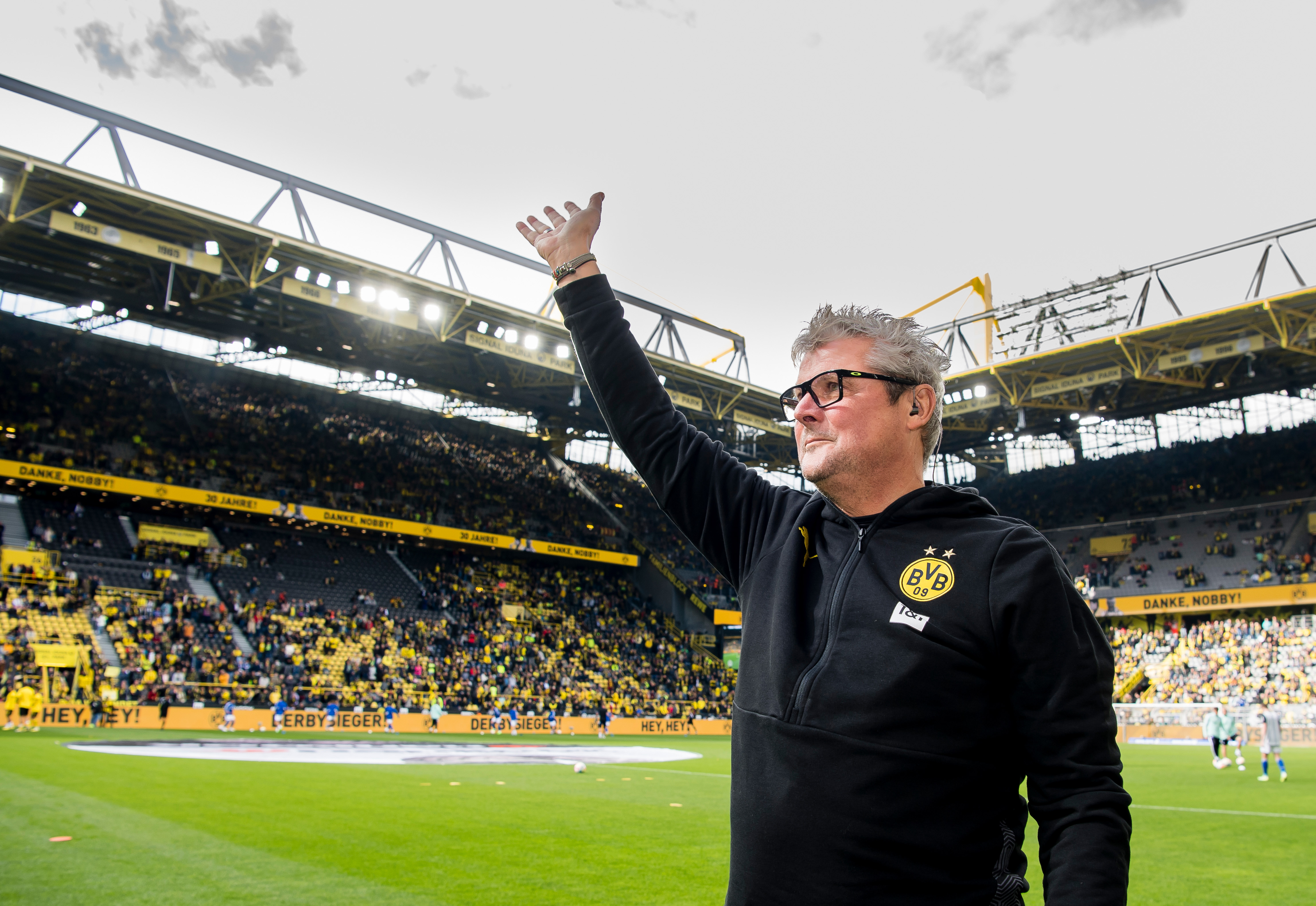 Foto eines Mannes, der auf dem Spielfeld eines gefüllten Fußballstadions die Hand in die Höhe reckt.