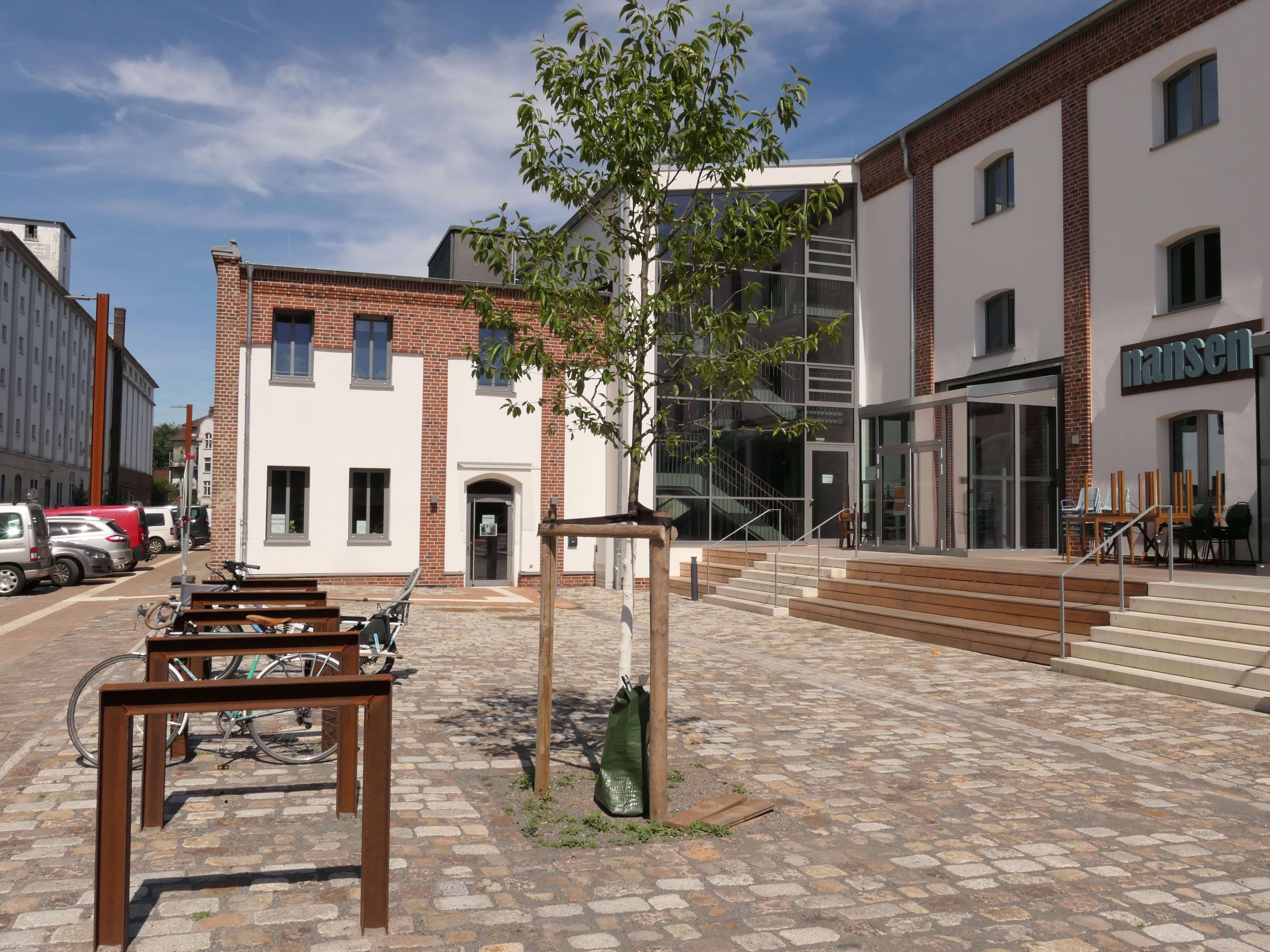 Fahrradbügel und ein junger Baum auf einem Kopfsteinpflasterplatz vor einem Gebäude mit teils sichtbarem, teils verputztem Mauerwerk