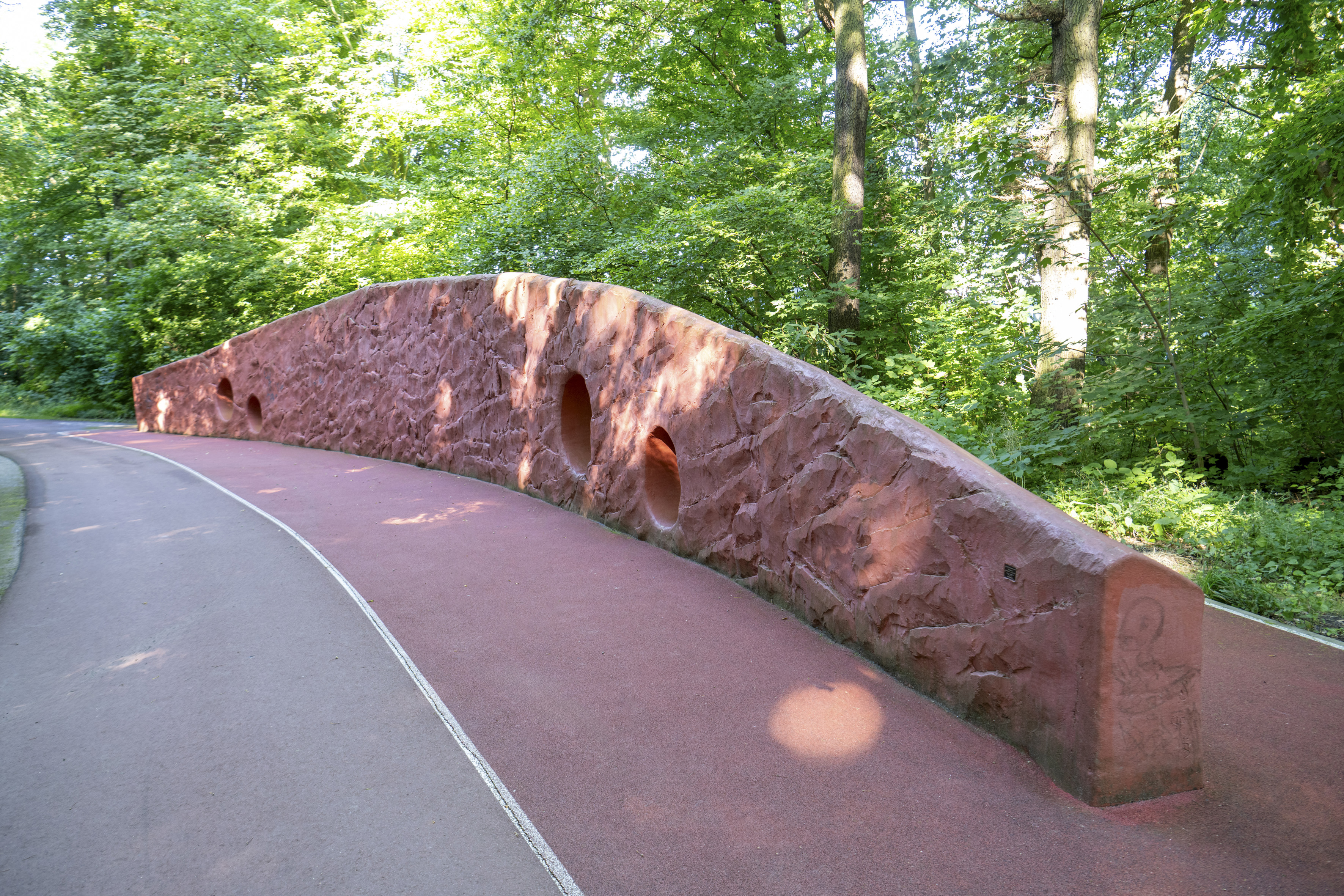 Eine rote, langgezogene Boulderwand entlang eines Weges, neben Bäumen. 