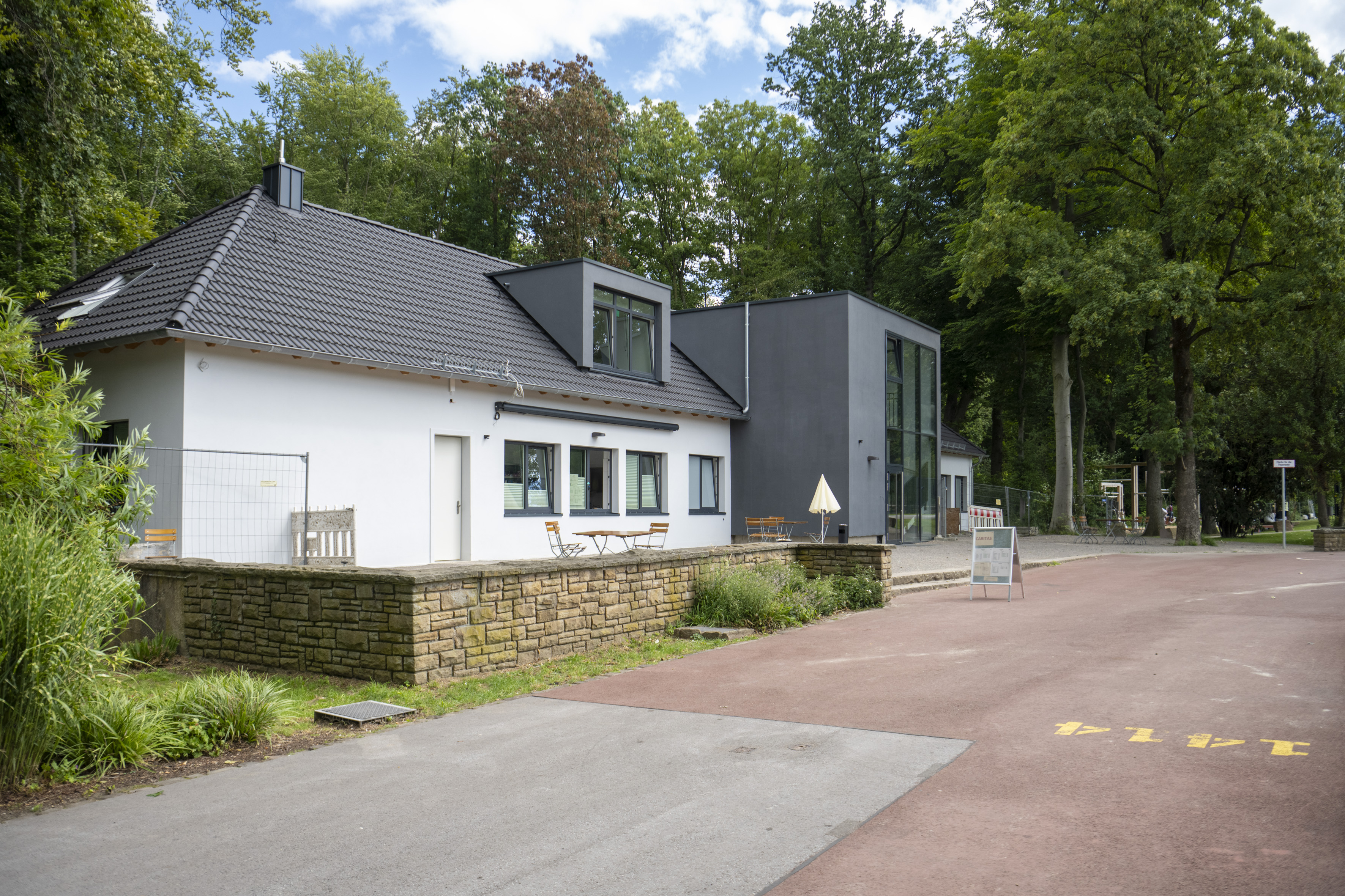 Ein zweistöckiges Gebäude mit Satteldach hinter einer flaschen Natursteinmauer, dahinter Wald.