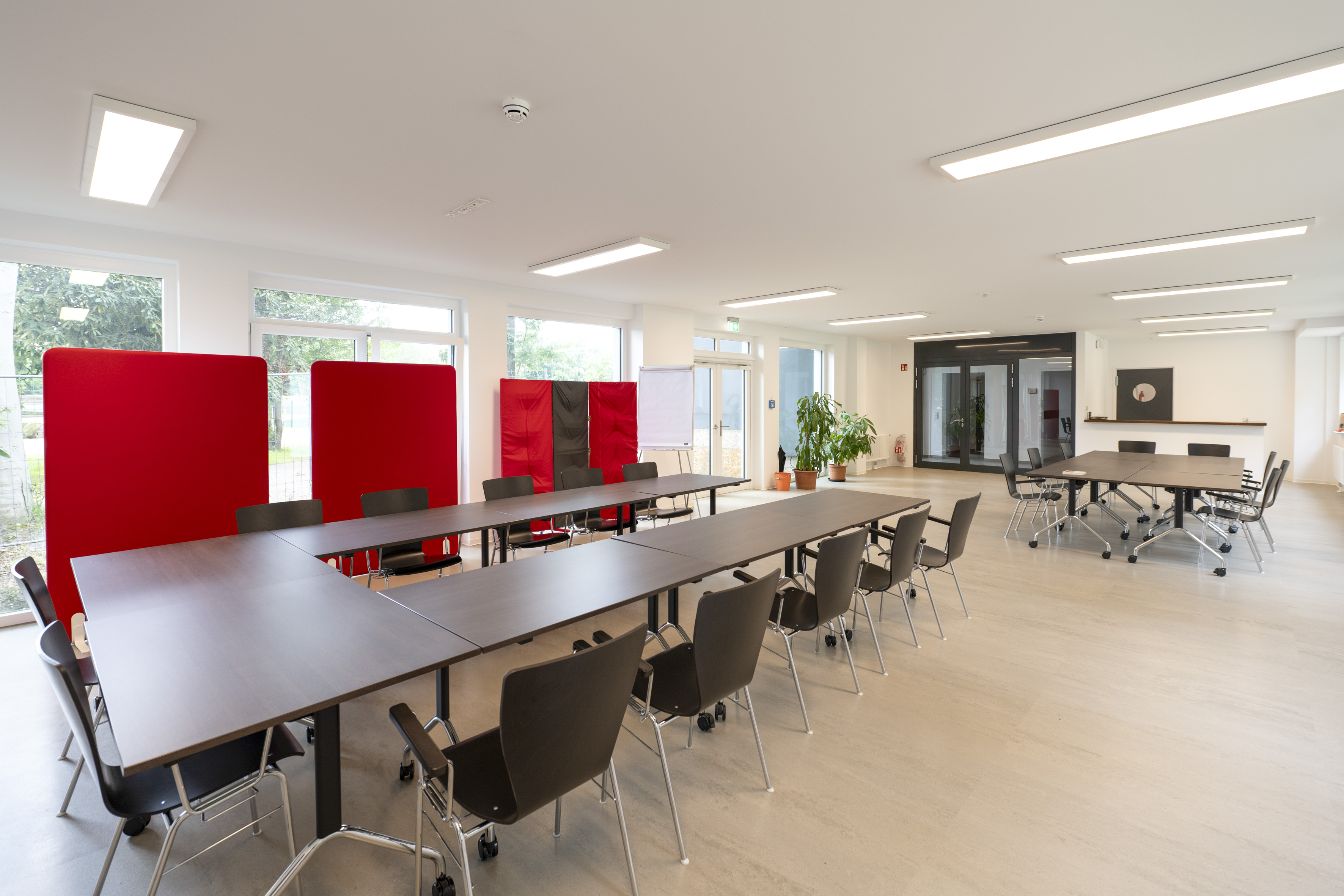 Heller großer Raum mit einer bestuhlten langen Tischreihe in der Mitte und roten Stellwänden vor großen Fensterflächen.