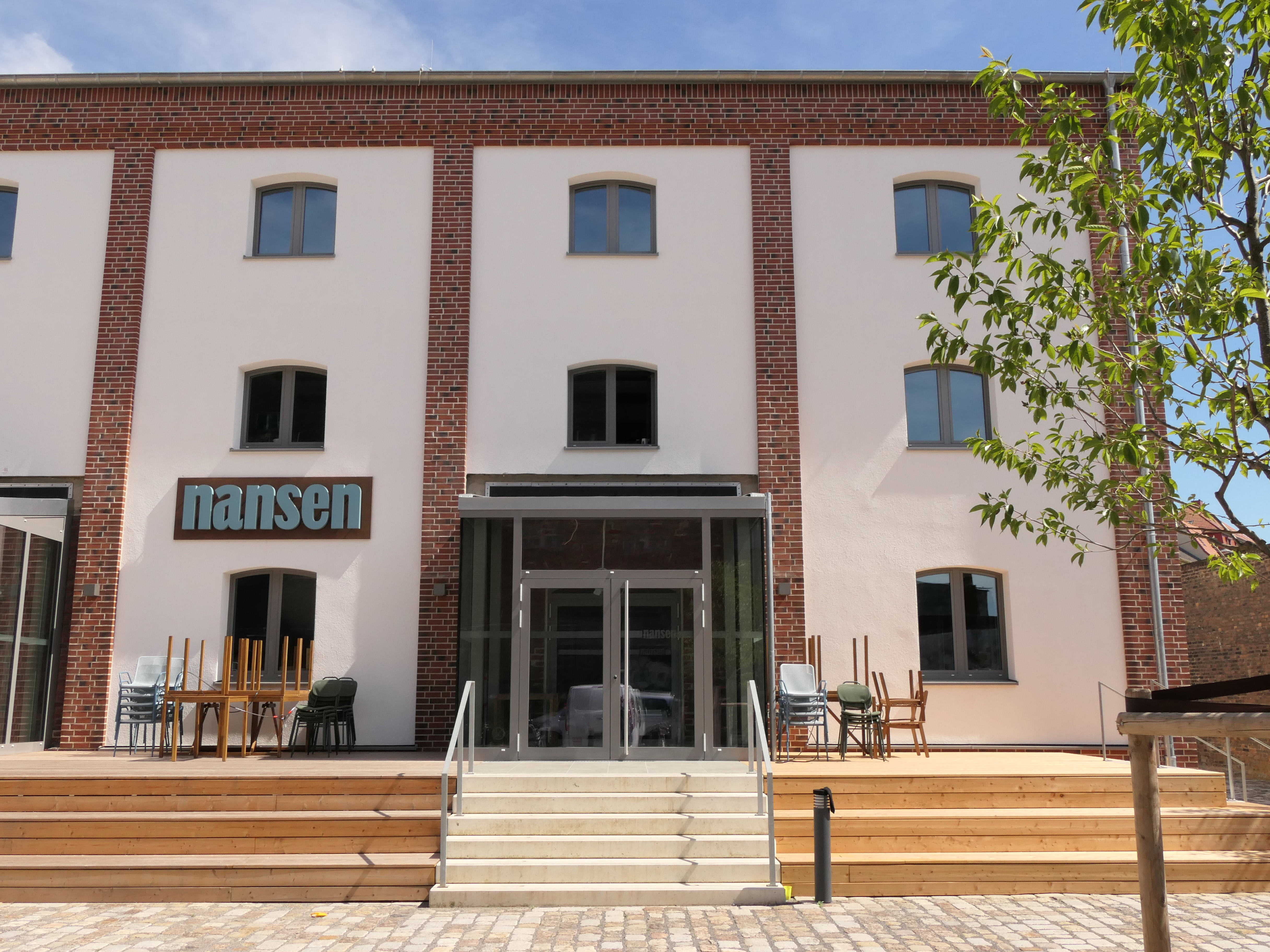 Frontalansicht eines Hauses mit vorgebauter Terrasse, Tischen und Stühlen und dem Schriftzug "nansen". 