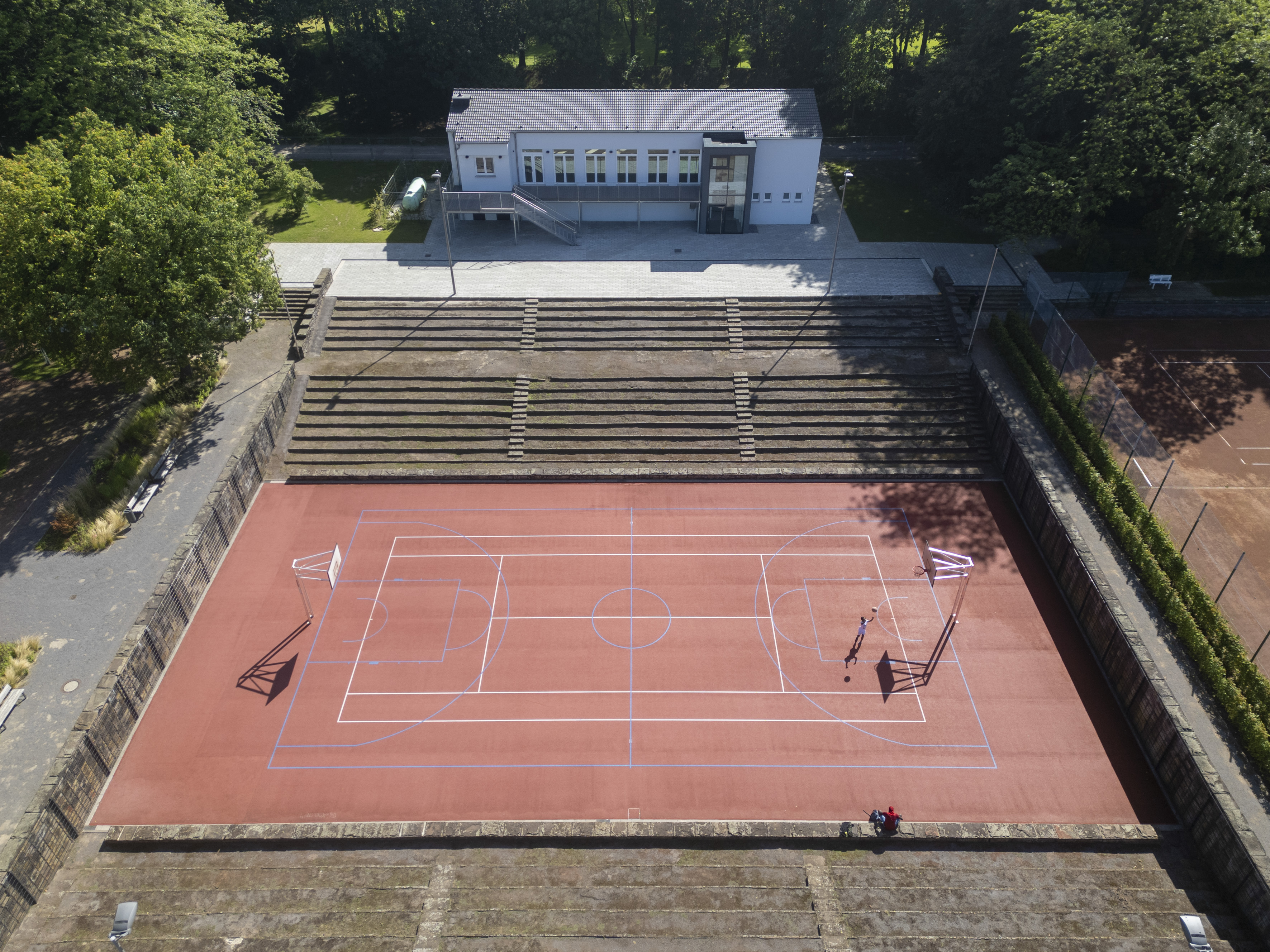 Drohnenaufnahme eines großen Basketballfeldes im Freien mit angrenzenden Tribünenstufen und einem Gebäude.