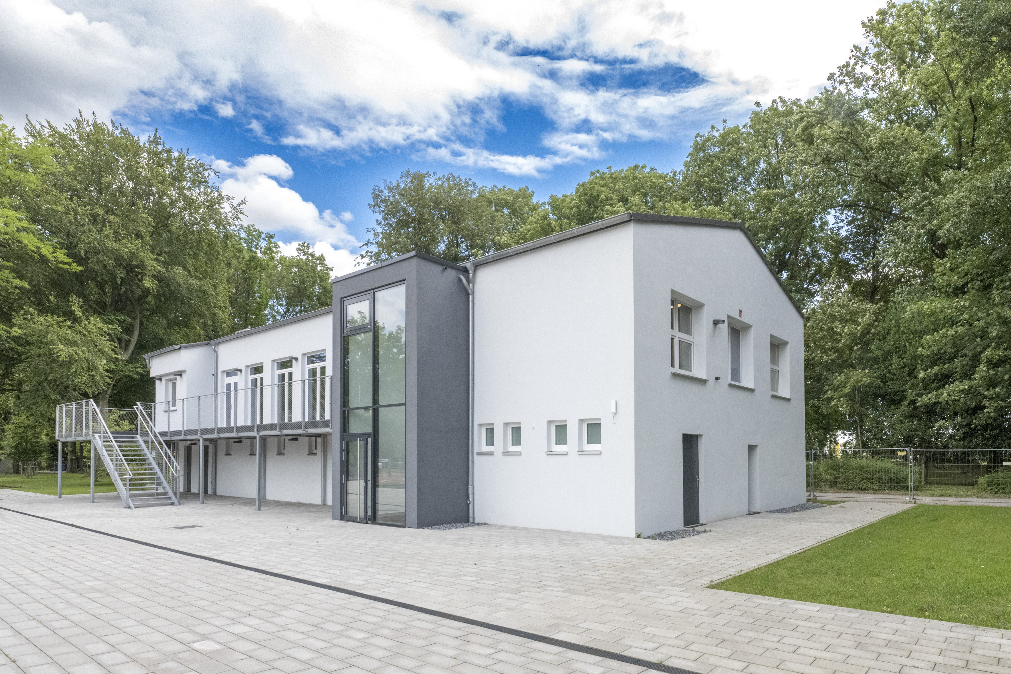 Ein zweistöckiges Gebäude mit Satteldach, Vorplatz und Bäumen im Hintergrund.