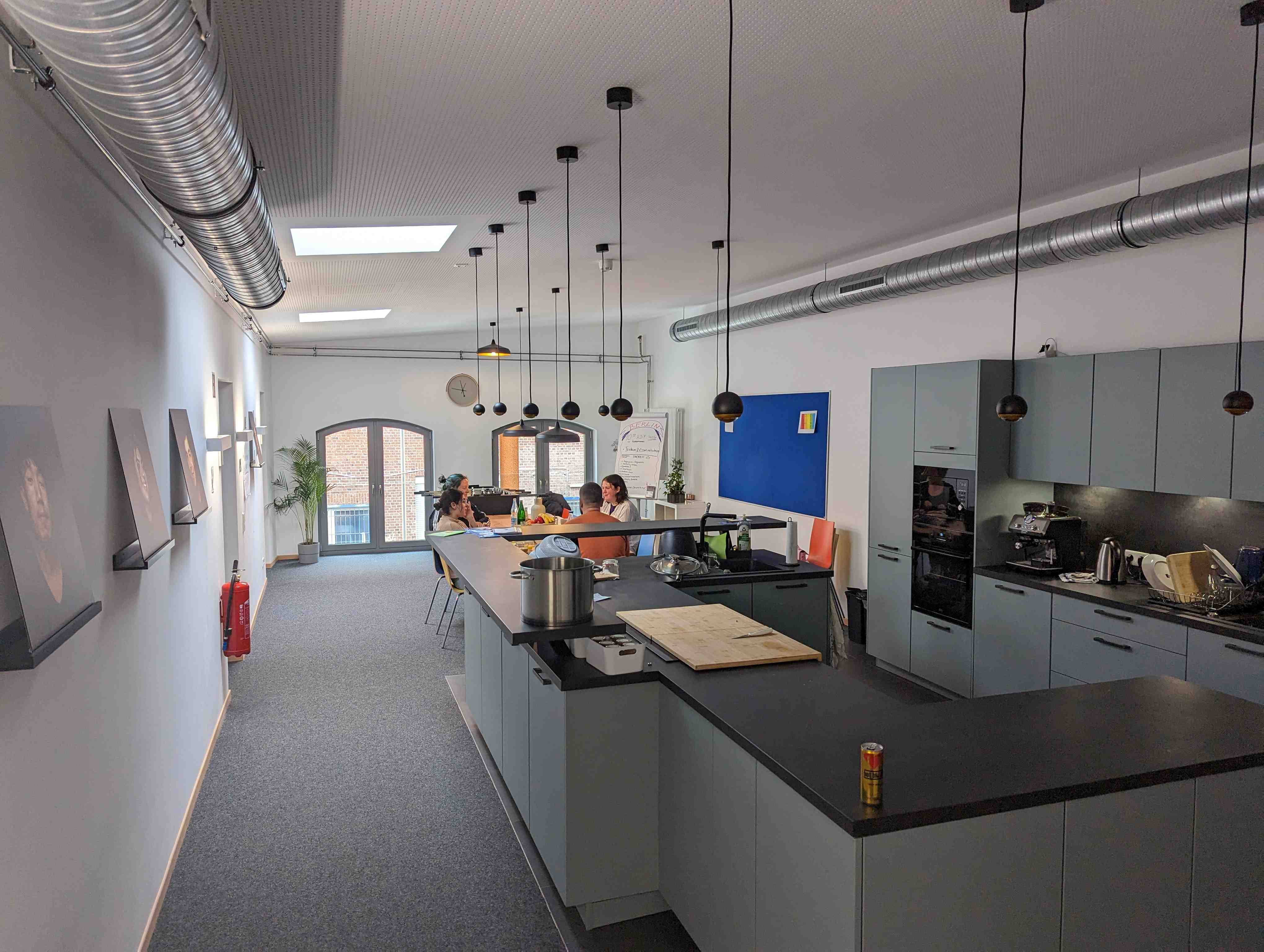 Ein Raum mit einer Küchenzeile im Vordergrund einer Gruppe Menschen an einem Tisch im Hiintergrund.