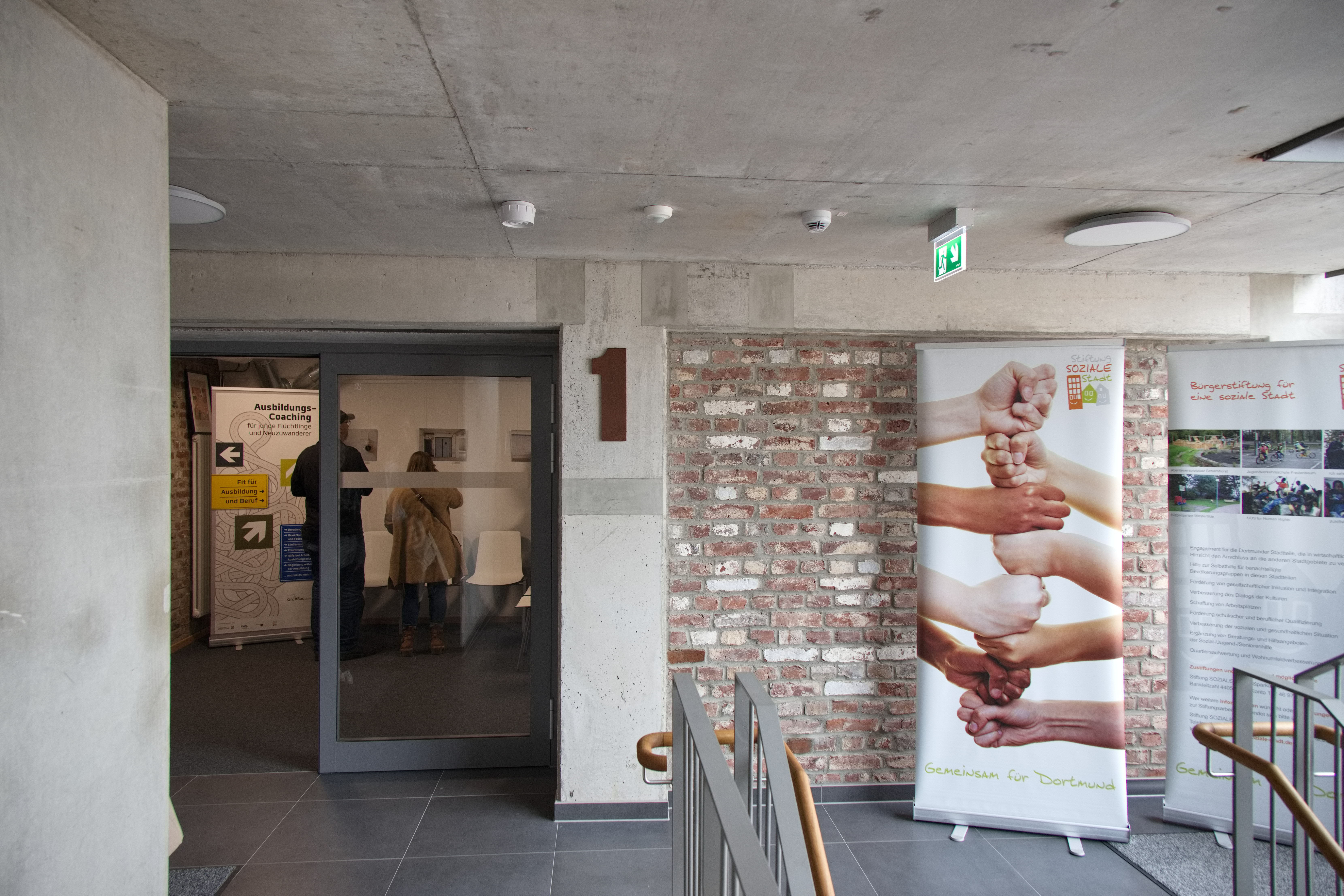 Blick aus einem Treppenhaus auf eine Glastür, daneben ein Roll-Up mit vielen Händen unterschiedlicher Hautfarbe.