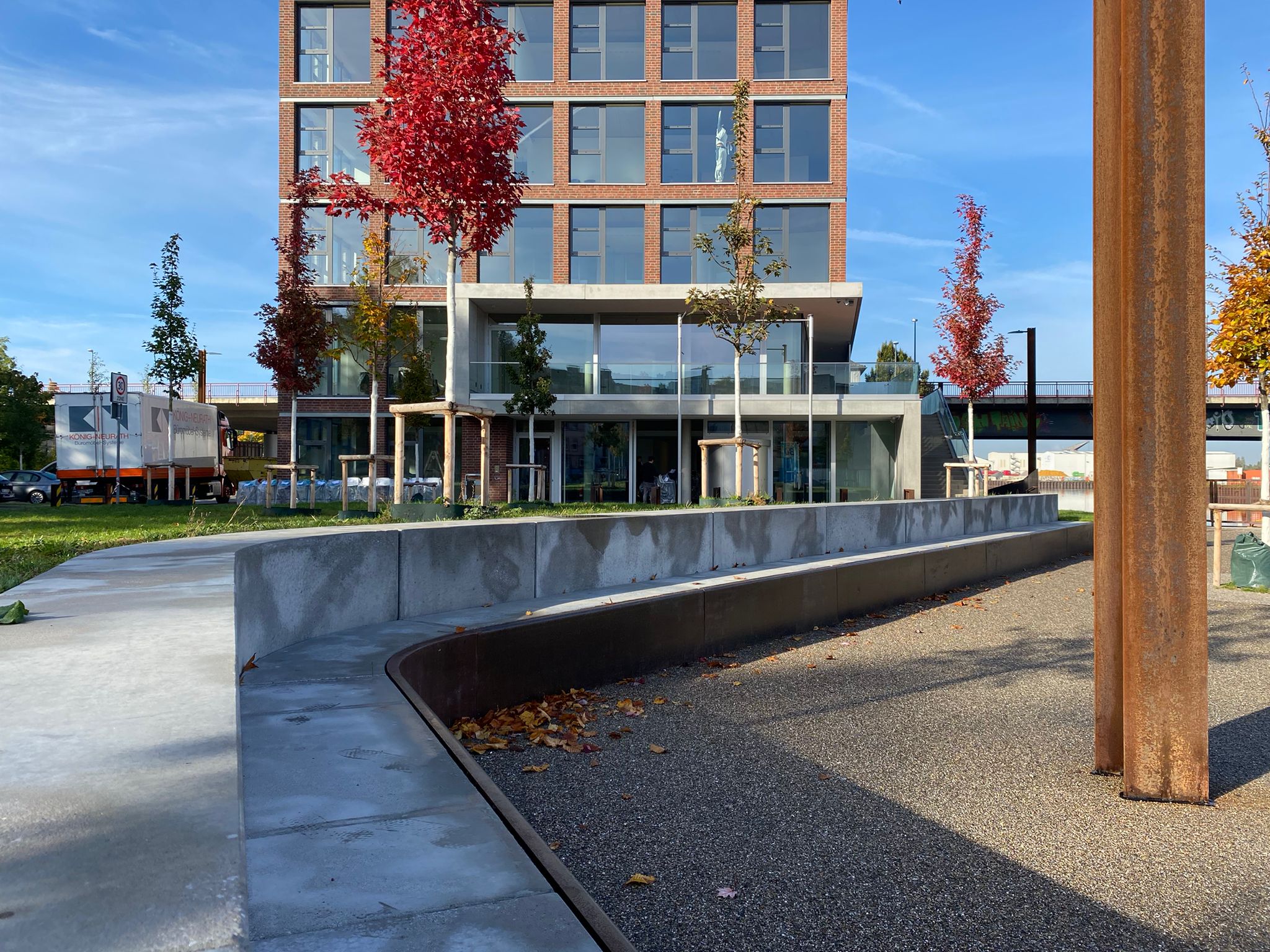 Langes Sitzelement aus Beton vor erhöhtem Rasendeck mit Bäumen und einem Neubau.