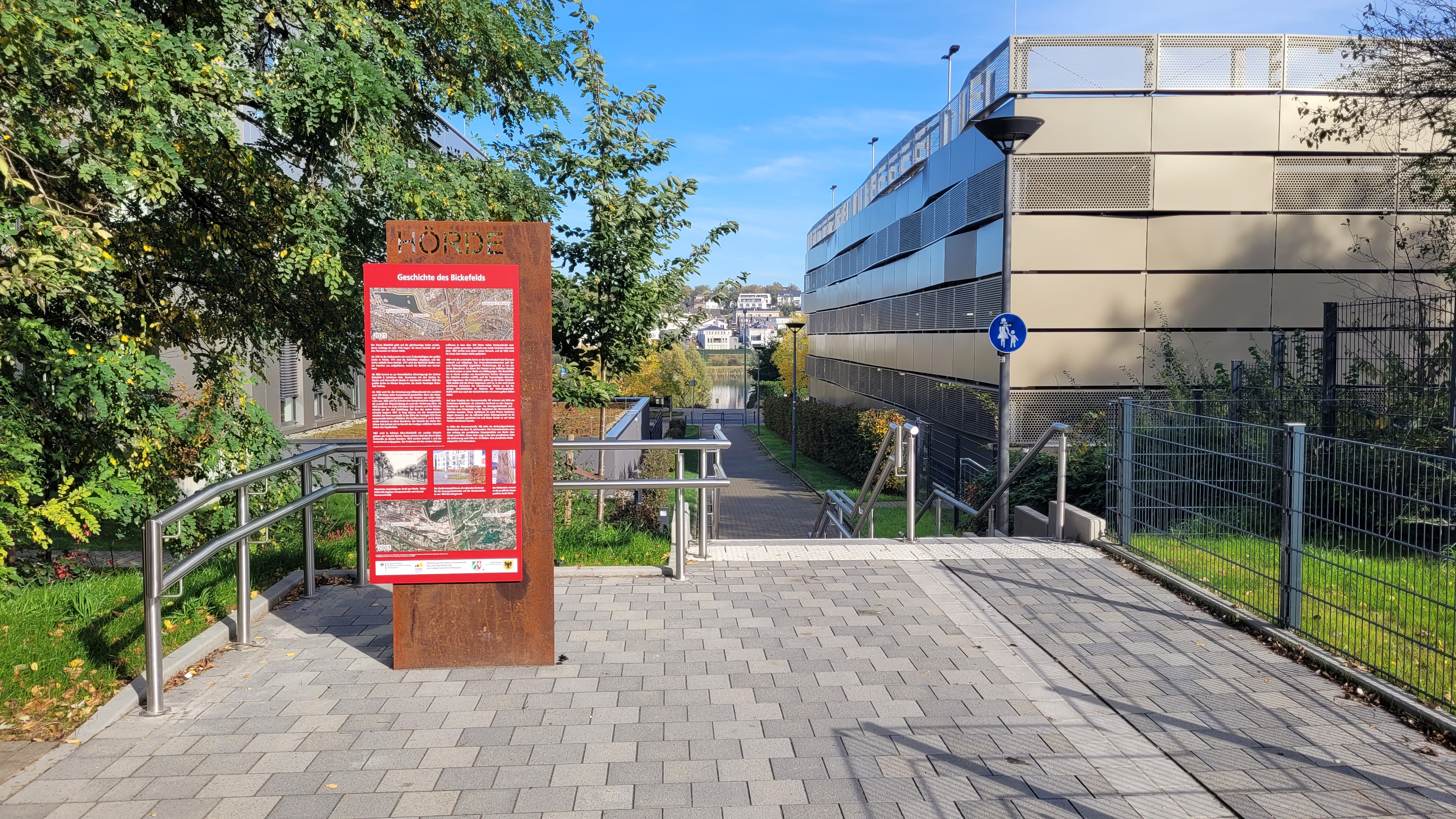 Ein Weg führt zwischen Häusern entlang über eine Treppe herab zu einem See, links steht eine Infostele.
