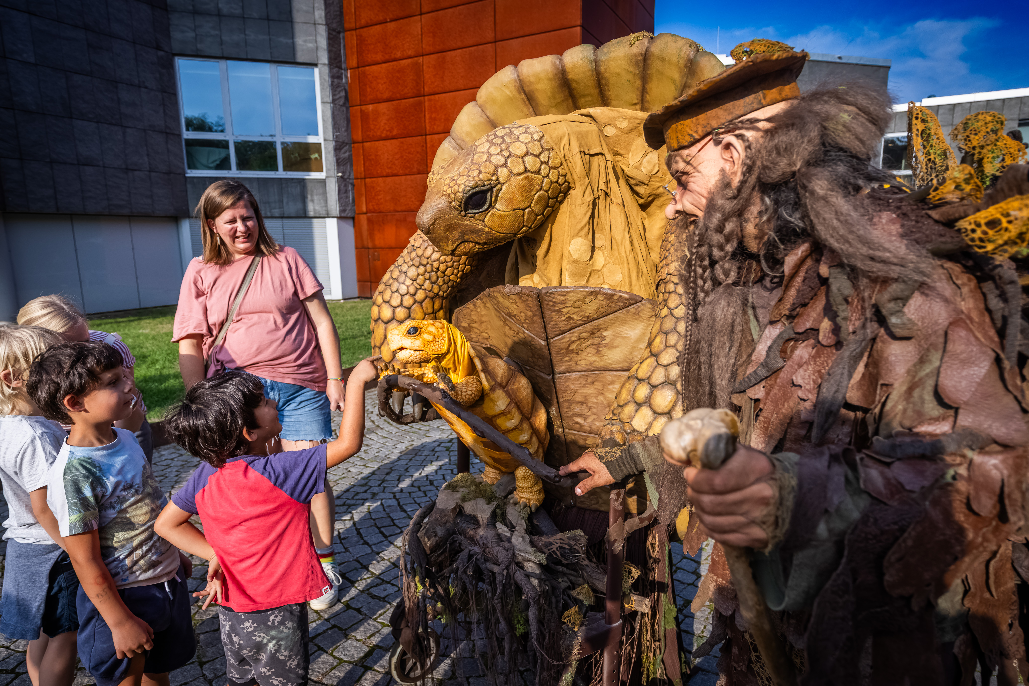 Walk-Act Großmutter Schildkröte: Mensch in riesigem Schildkrötenkostüm mit Besucher*innen vor dem Naturmuseum und vor blauem Himmel
