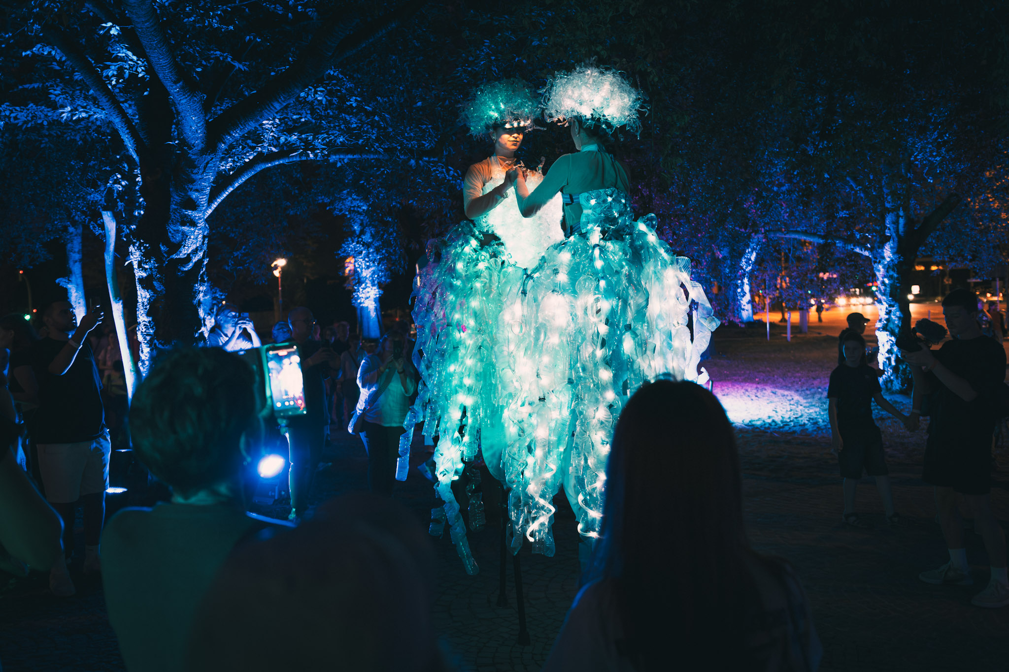 Zwei Stelzenläuferinnen in beleuchteten schwingenden Kleidern, die sich ansehen und an den Händen halten. Drumherum stehen Personen, die zugucken.