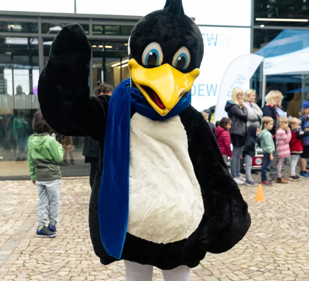 Zu sehen ist das DEW21-Maskottchen Pido. Pido ist ein Pinguin und steht winkend vor dem DEW21 Servicecenter. Im Hintergrund sind Kinder zu sehen.