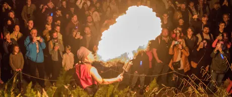 Zu sehen ist ein Feuerspucker und eine große Flamme. Im Hintergrund stehen sehr viele Besucher*innen.