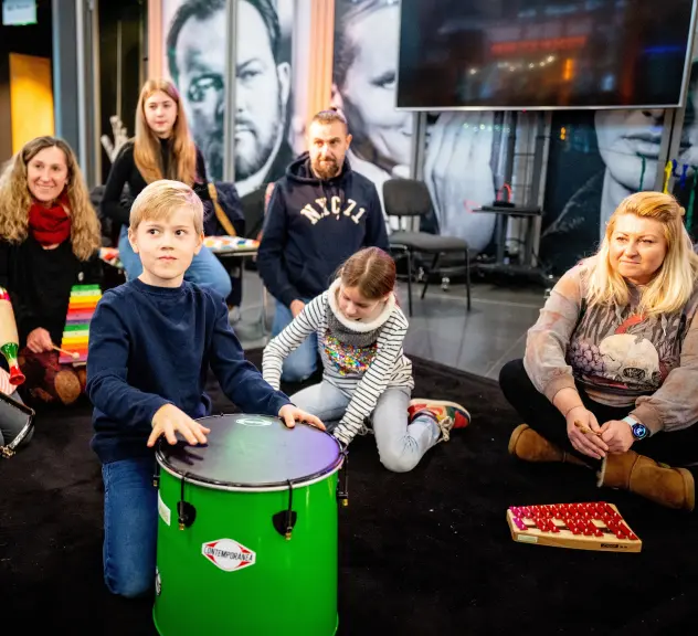 Drei Erwachsene und vier Kinder sitzen mit verschiedenen Instrumenten auf dem Boden. Unter den Instrumenten sind eine Rassel, eine Trommel und ein Xylophon.