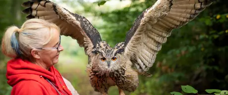 Zu sehen ist eine Frau mit einer großen schwarz-braunen Eule auf dem Arm. Die Eule hat ausgebreitete Flügel.