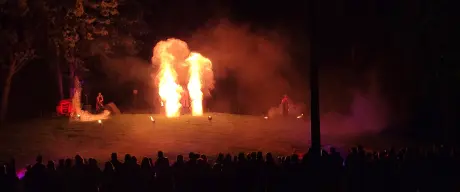 Mittig sind zwei Feuer-Fontänen zu sehen, auf dem Boden stehen kleine Fackeln. Vorne stehen viele Zuschauer*innen.