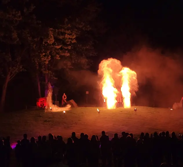 Mittig sind zwei Feuer-Fontänen zu sehen, auf dem Boden stehen kleine Fackeln. Links und rechts steht jeweils eine Person, die ebenfalls Feuer machen. Vorne stehen viele Zuschauer*innen.