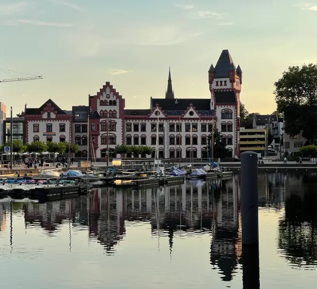 Zu sehen ist die rot-weiße Hörder Burg bei Einbruch des Abends. Sie spiegelt sich auf der Hafen-Seite im Phönix See. Auf dem Wasser befinden sich einige kleine Boote sowie Tretboote.