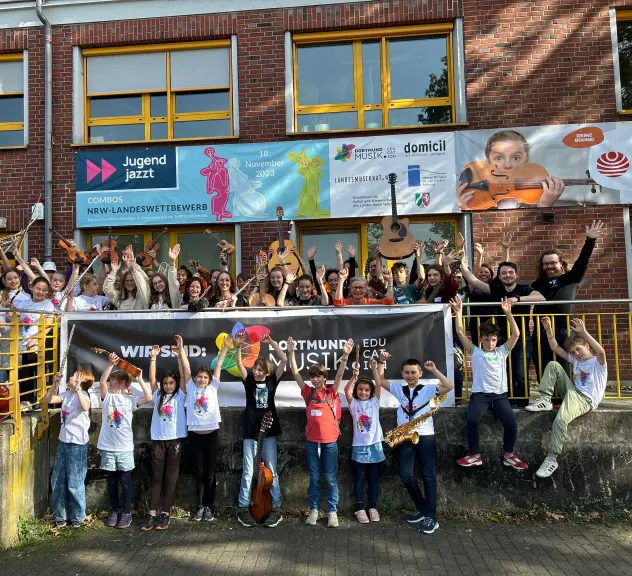 Gruppenfoto der Schüler*innen von Dortmund Musik vor dem Gebäude der Musikschule. Kinder verschiedenen Alters recken die Hände und teils ihre Instrumente in die Höhe, lächeln und gucken in die Kamera.