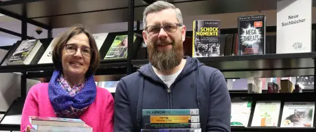 Ein Mann und eine Frau stehen mit dem Rücken zu einem Bücherregal in der Bibliothek und lächeln in die Kamera.