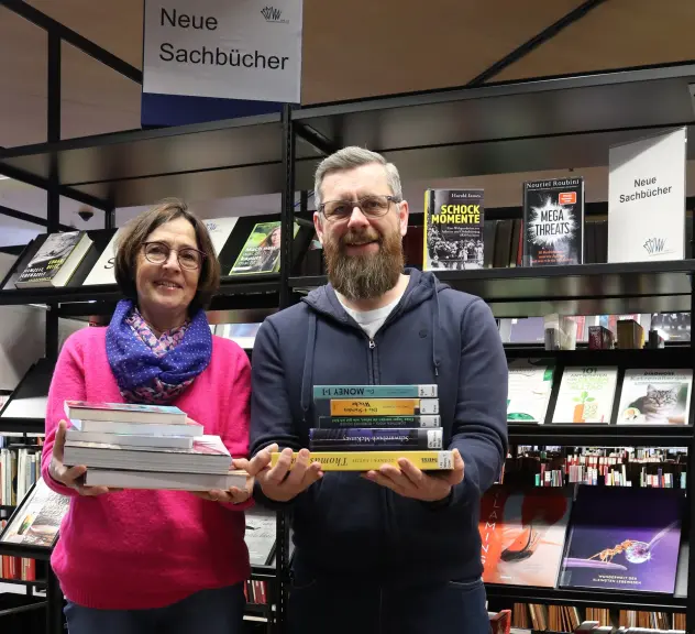 Ein Mann und eine Frau stehen mit dem Rücken zu einem Bücherregal in der Bibliothek und lächeln in die Kamera. Über dem Regal hinter ihnen steht "Neue Sachbücher". Beide halten Bücherstapel in den Händen..