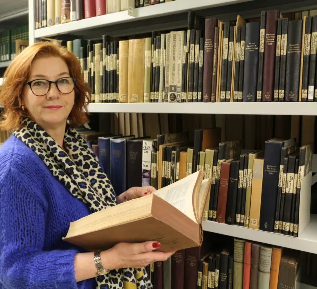 Foto von Heike Fonteyne, die vor einem Bücherregal mit alten Büchern steht. In den Händen hält sie ein aufgeschlagenes altes Buch und blickt dabei freundlich in die Kamera.