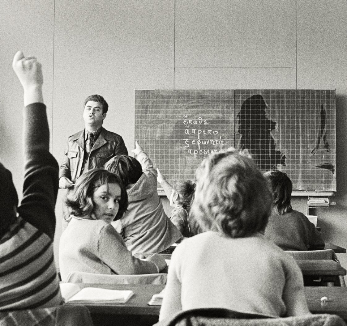 Schwarz-weiß Aufnahme einer Schulklasse von hinten. Vorne ist der Lehrer zu sehen, an der Tafel stehen griechische Buchstaben. Ein Mädchen dreht sich um und blickt in die Kamera.